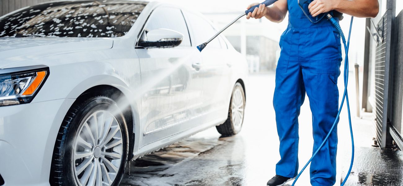 Professional washer in blue uniform washing luxury car with water gun on an open air car wash. Close up photo.